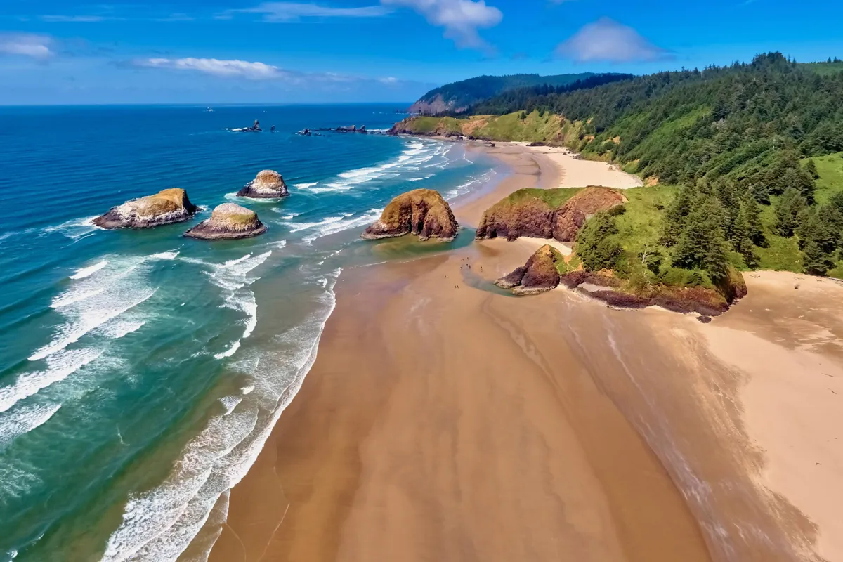 A tranquil view of sandy beach and dramatic rock formations, with green hills in the background and gentle waves rolling in. The scene suggests opportunities for outdoor relaxation, picnicking, and enjoying nature just moments from the motel.