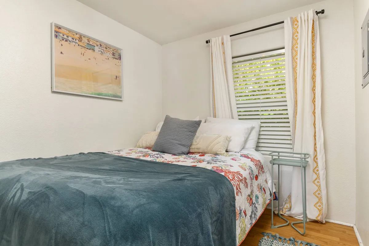 A cozy bedroom featuring a full-sized bed adorned with floral bedding. The room is softly lit by natural daylight filtering through curtains by the window, and tasteful artwork hangs on the wall to create a serene atmosphere.
