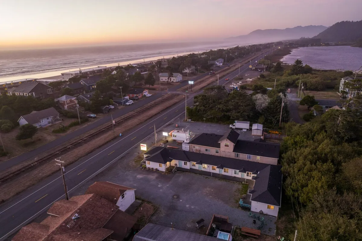 This image provides a broad view of the motel's surroundings, featuring the ocean, a road, local buildings, and distant mountains. The scene captures a serene sunset, warm skies, and a hint of community through adjacent buildings, evoking the atmosphere of a welcoming coastal gathering place.