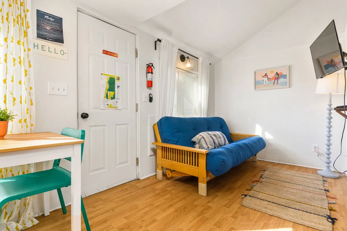 The image showcases a cozy living space with a futon sofa, a small dining table set for two, a window with light curtains filtering in natural light, a television for entertainment, and charming artwork on the wall. The layout encourages relaxation and socializing in an inviting, compact setting.