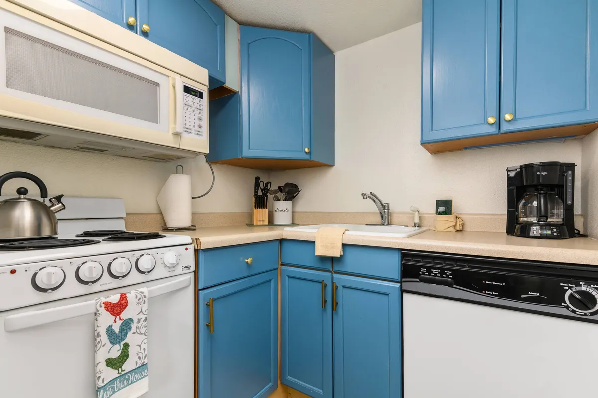 A cozy, homey kitchen with blue cabinetry, white appliances, and thoughtfully arranged kitchen accessories. The kitchen is compact but functional, suggesting an inviting space for preparing meals, baking, or socializing with friends and family.
