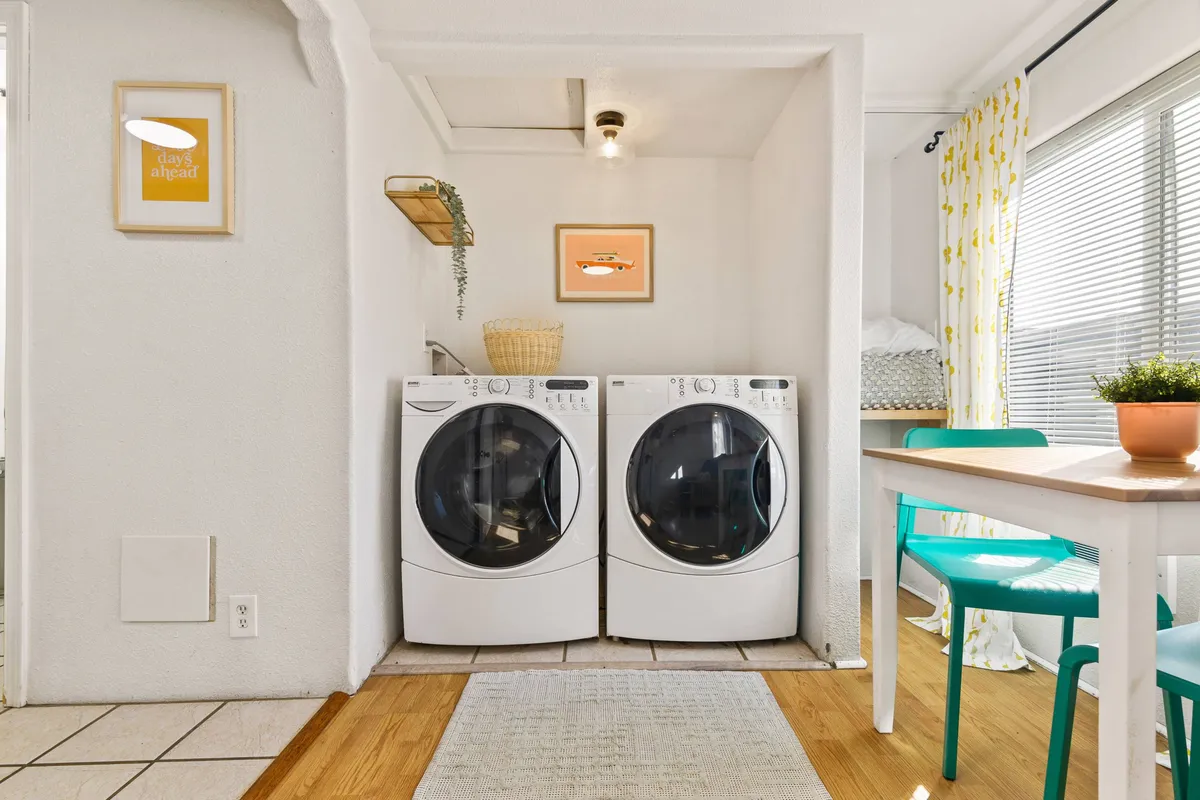 This image showcases a bright and cheerful interior space featuring a washer and dryer, a small table with a plant, and decorative wall art. The mood is practical and inviting, suggesting a comfortable place to take care of basic needs while relaxing.